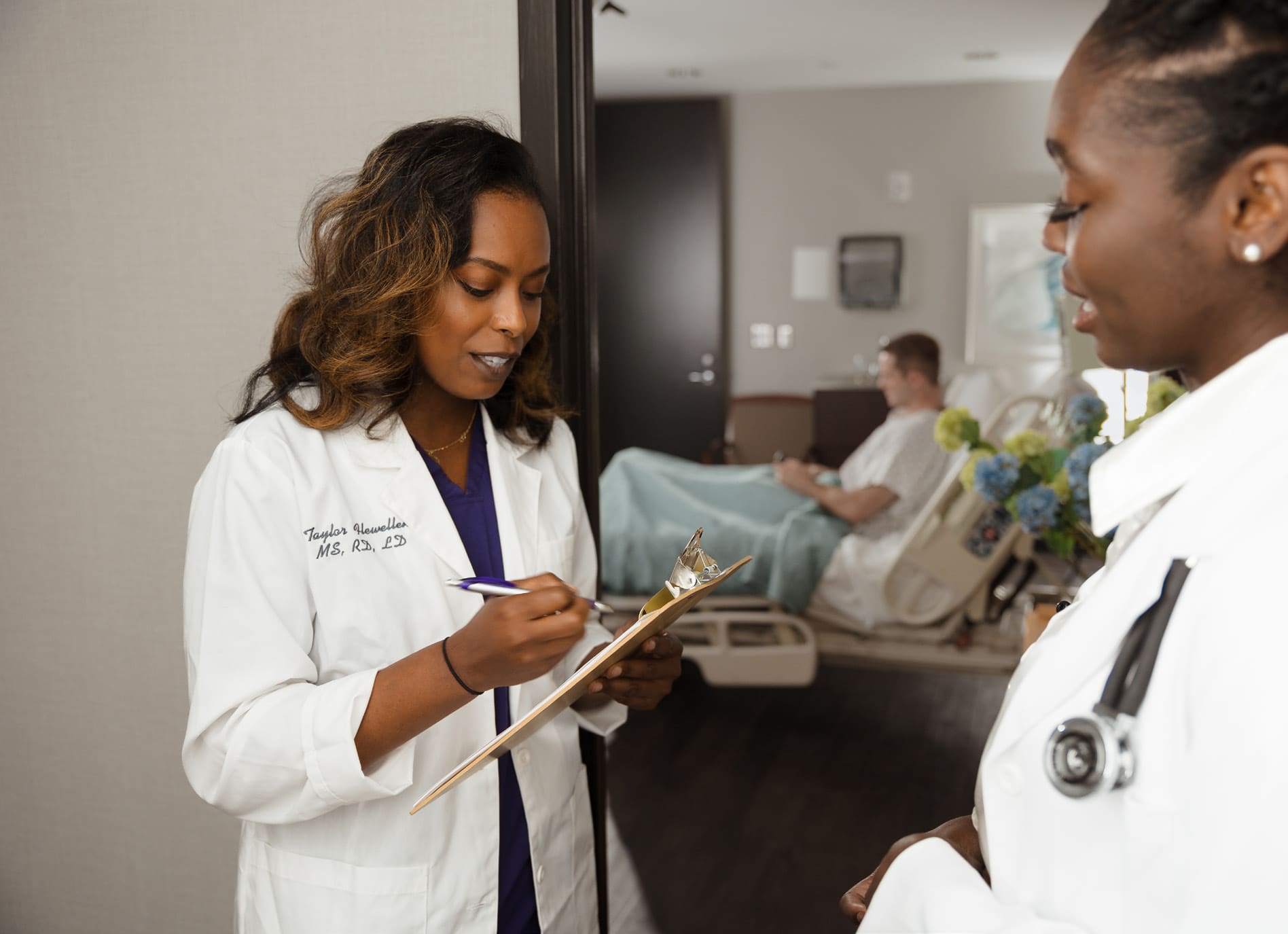 Two Morrison healthcare professionals in white lab coats discussing a patient's chart in a hospital room, with a patient lying in a hospital bed in the background.